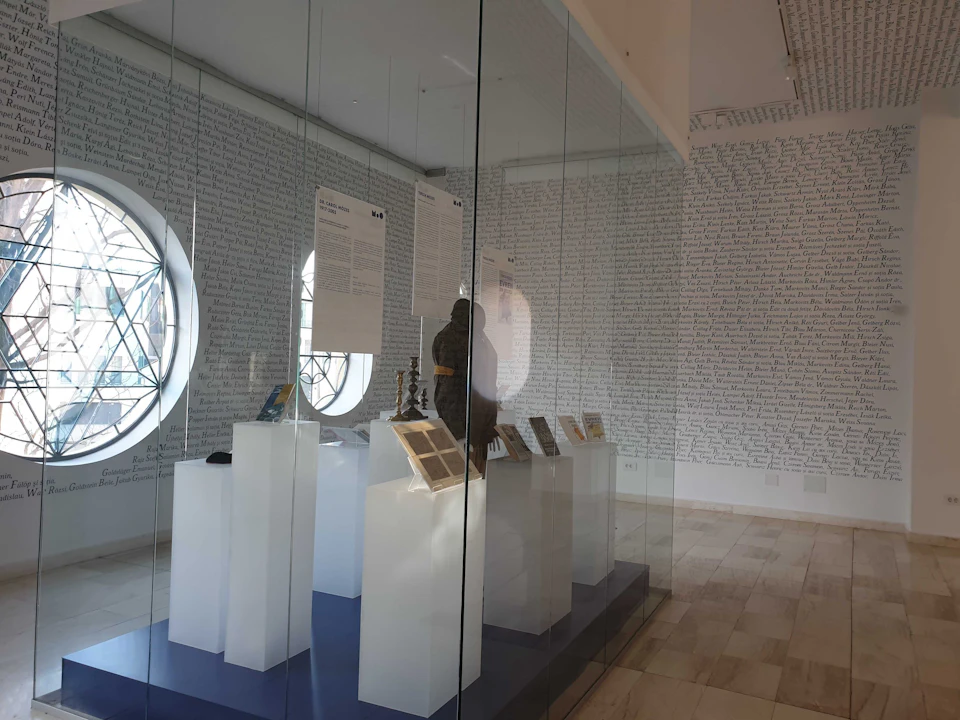 Exhibit with artifacts on white pedestals behind glass display, with two round windows and walls covered in handwritten text.