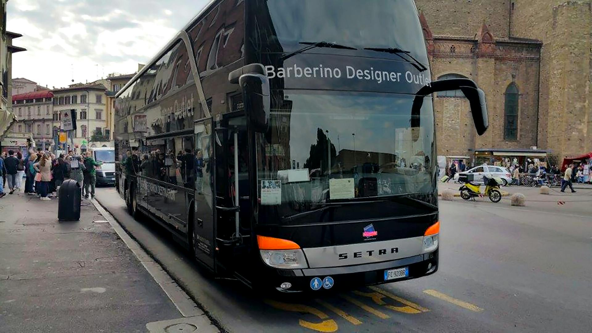 Un autobús negre de dos pisos amb l'etiqueta "Barberino Designer Outlet" està aparcat en un carrer molt concorregut prop d'un gran edifici històric.