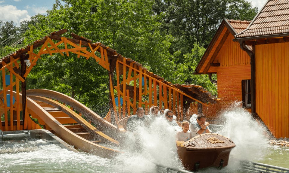 People on a wooden log flume ride splash into water, surrounded by lush greenery and a wooden structure.
