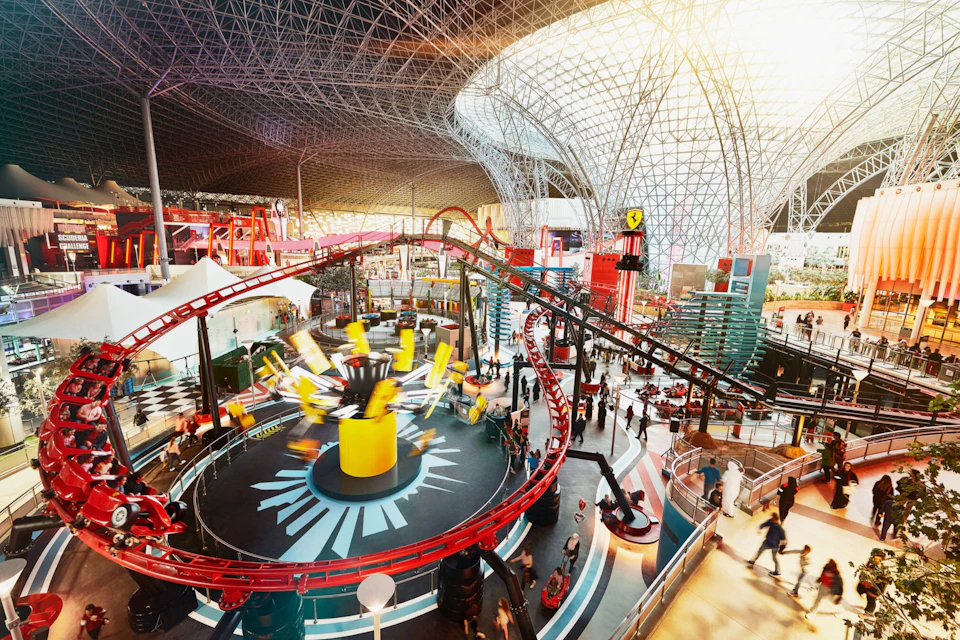 Indoor amusement park with a rollercoaster and spinning ride under a large glass and steel dome structure.