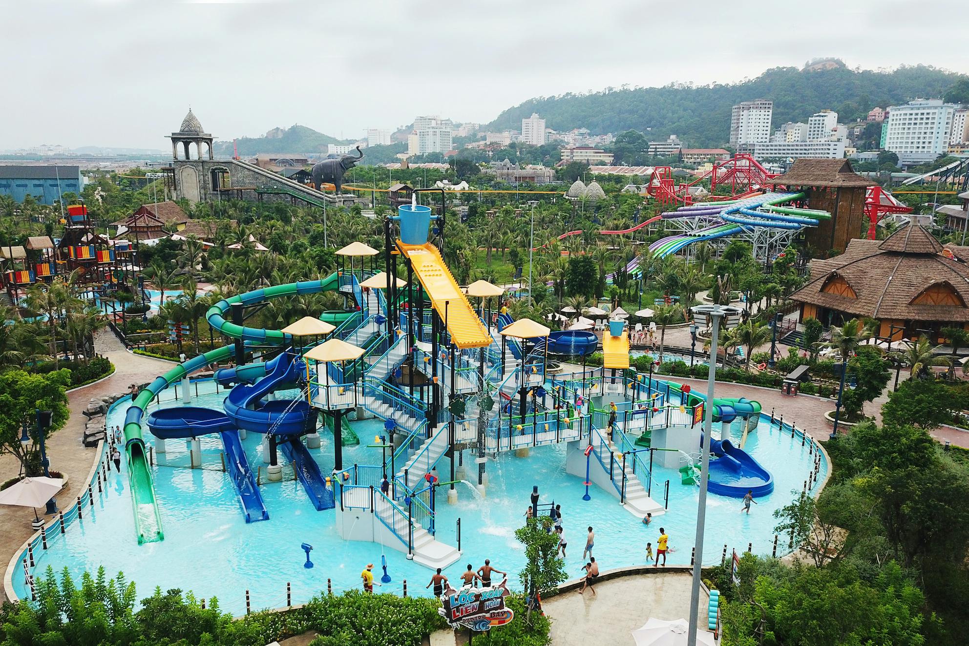 A water park with slides and pools, surrounded by greenery and buildings in the background, on a cloudy day.