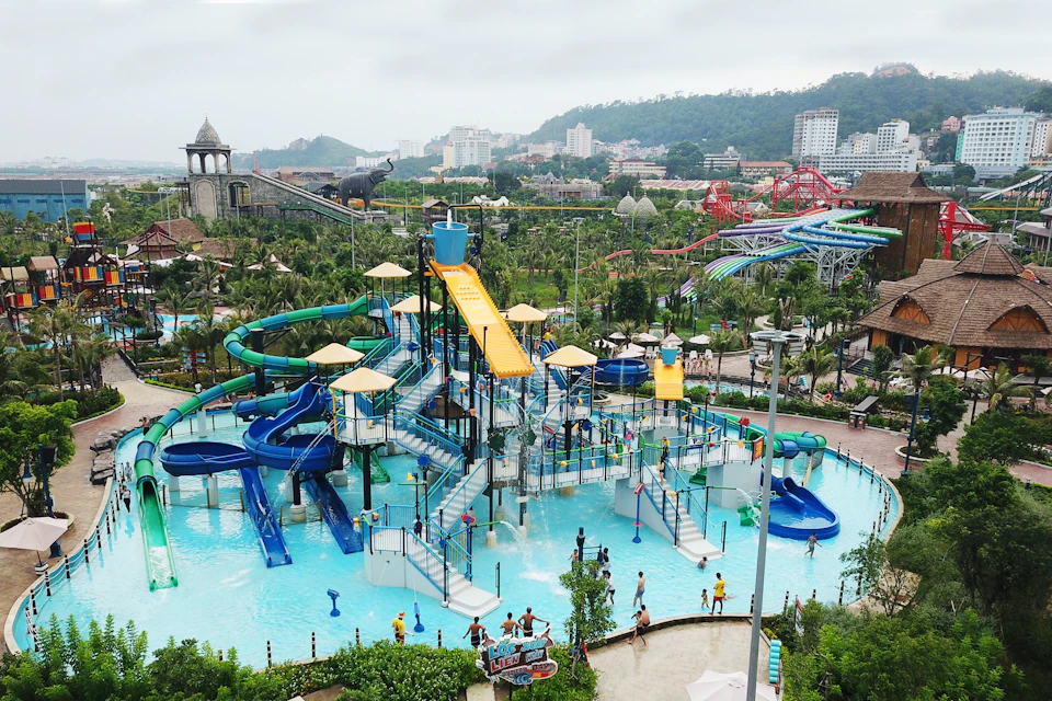 A water park with slides and pools, surrounded by greenery and buildings in the background, on a cloudy day.