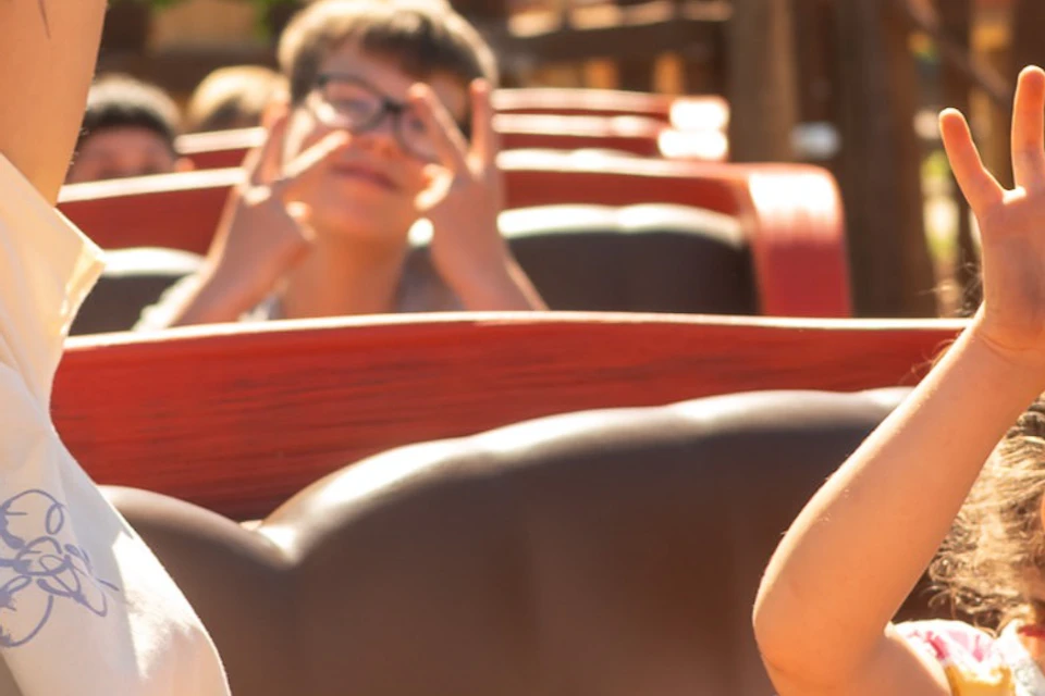 A woman and child on a barrel-themed amusement ride, raising their hands in enjoyment, with a rustic setting in the background.