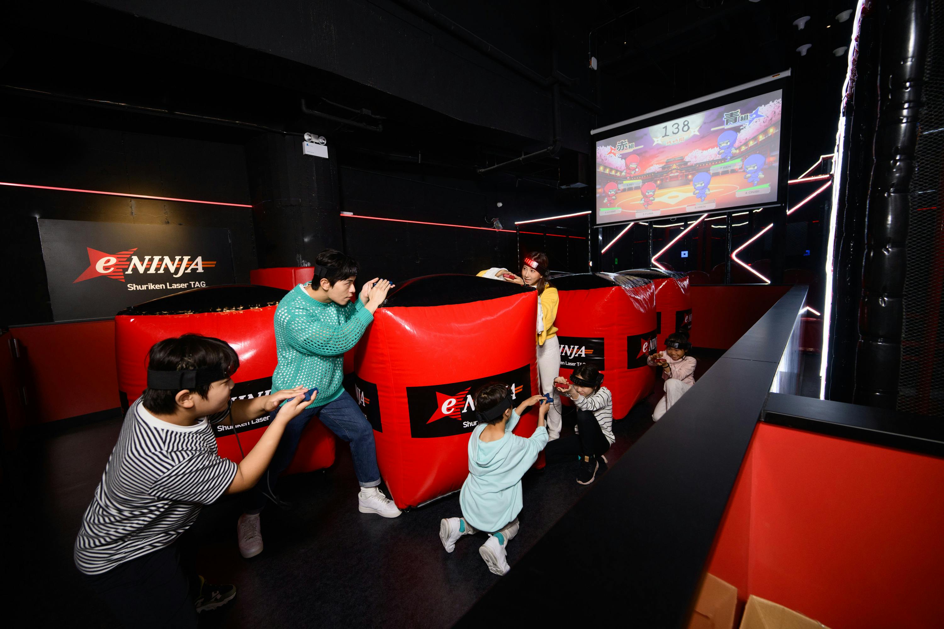 Children playing laser tag in a dark arena with red barriers, a screen displaying a game, and neon lighting accents.