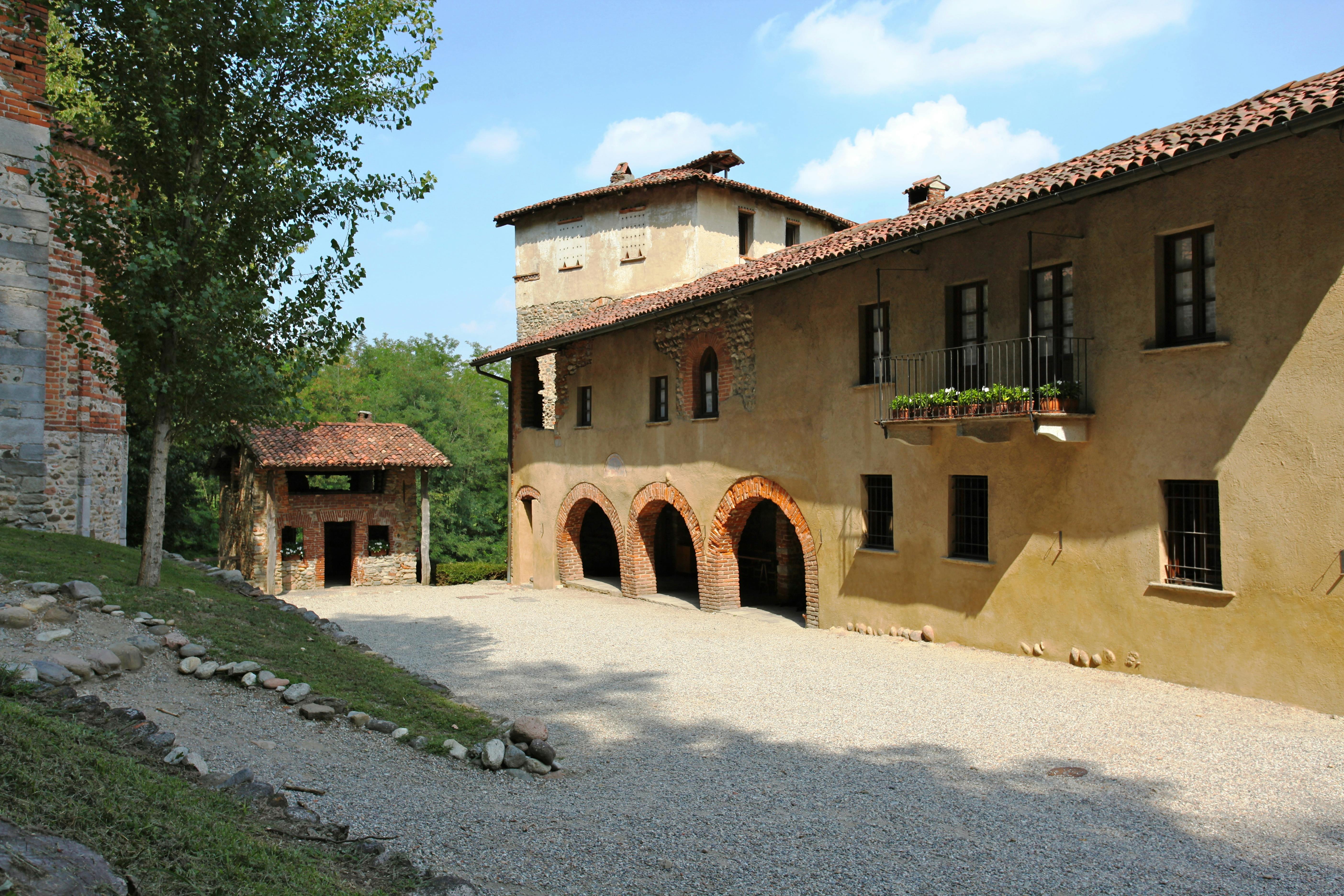 Stone building with arched entrances, a gravel courtyard, a smaller structure on the left, and trees in the background under a blue sky.