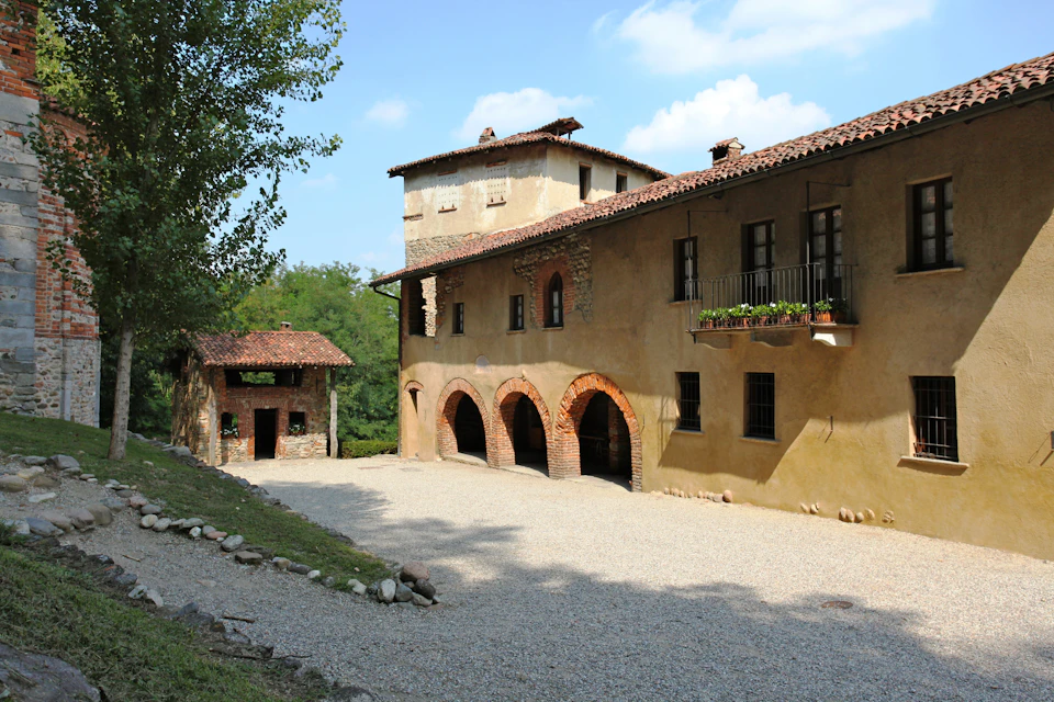 Stone building with arched entrances, a gravel courtyard, a smaller structure on the left, and trees in the background under a blue sky.