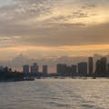 Solnedgångens omfamning över Miamis skyline: höga byggnader, moln, en touch av gul sol och avlägsna båtar på lugnt vatten.