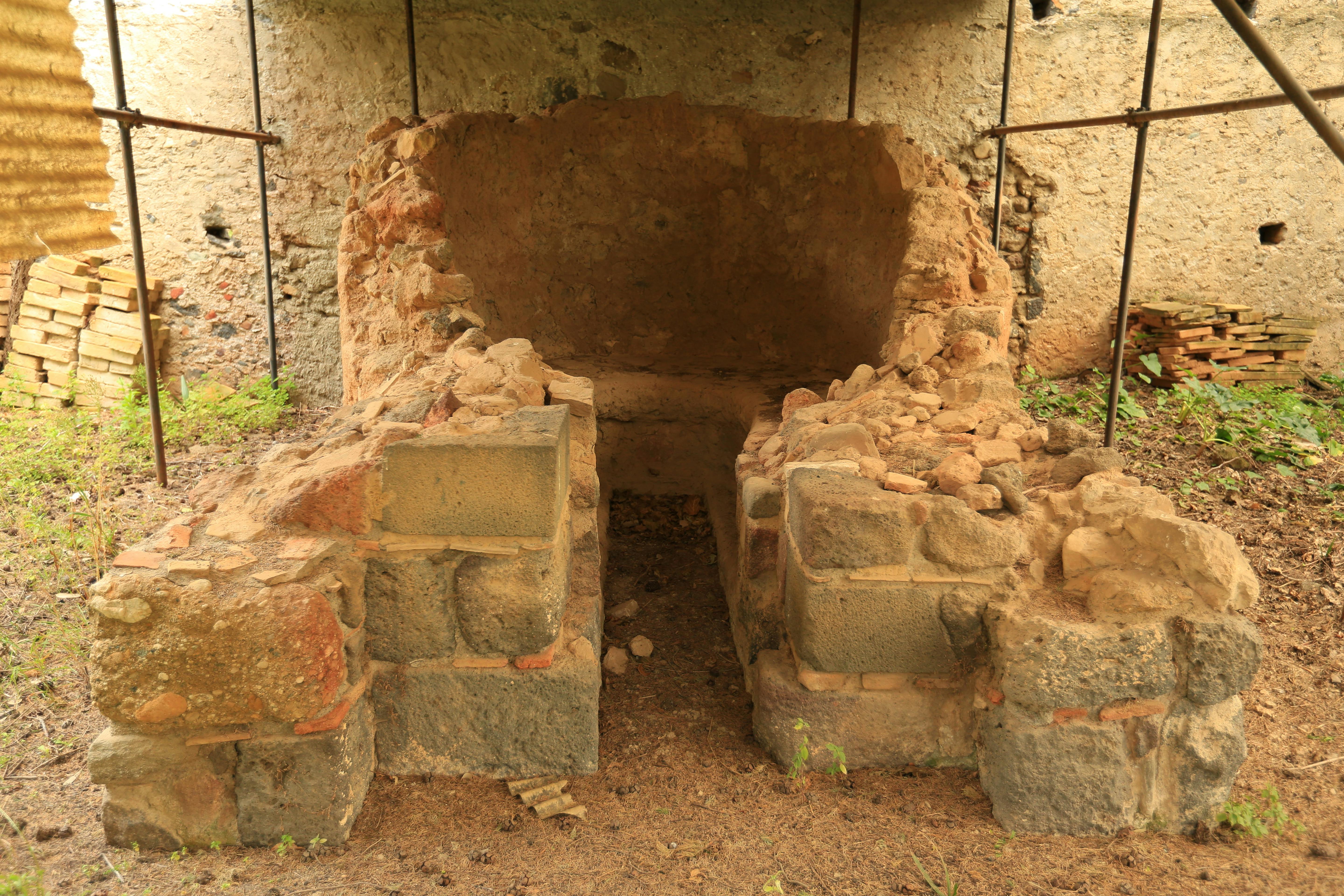 Ancient stone oven with archway, partially crumbled, surrounded by earth and small plants in a rustic outdoor setting.