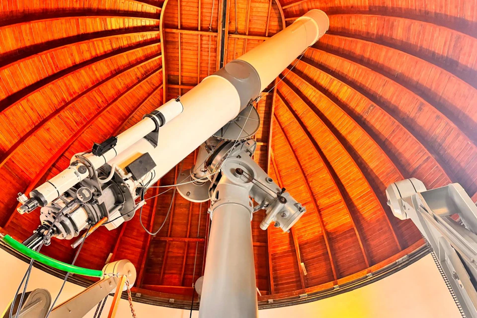Large telescope inside a wooden-domed observatory ceiling with visible mechanical components and cables.