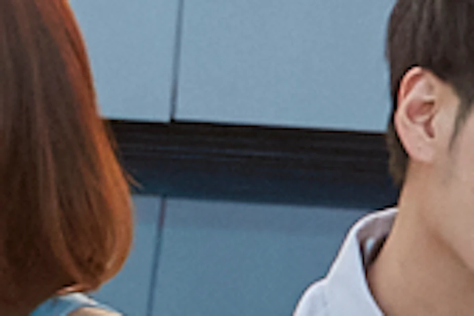 A man and woman sit on a glass floor rooftop with a cityscape and sunset in the background.