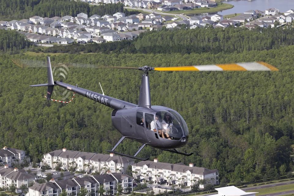 A black helicopter with two visible passengers flies over a residential area and a dense forest.