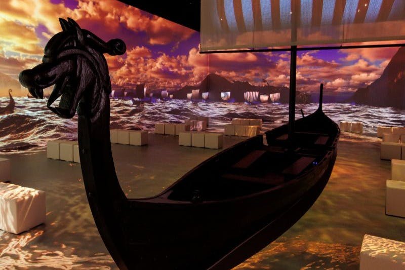 Un barco oscuro de estilo vikingo expuesto en una sala con una proyección de fondo de mar y cielo y asientos en forma de cubo blanco.
