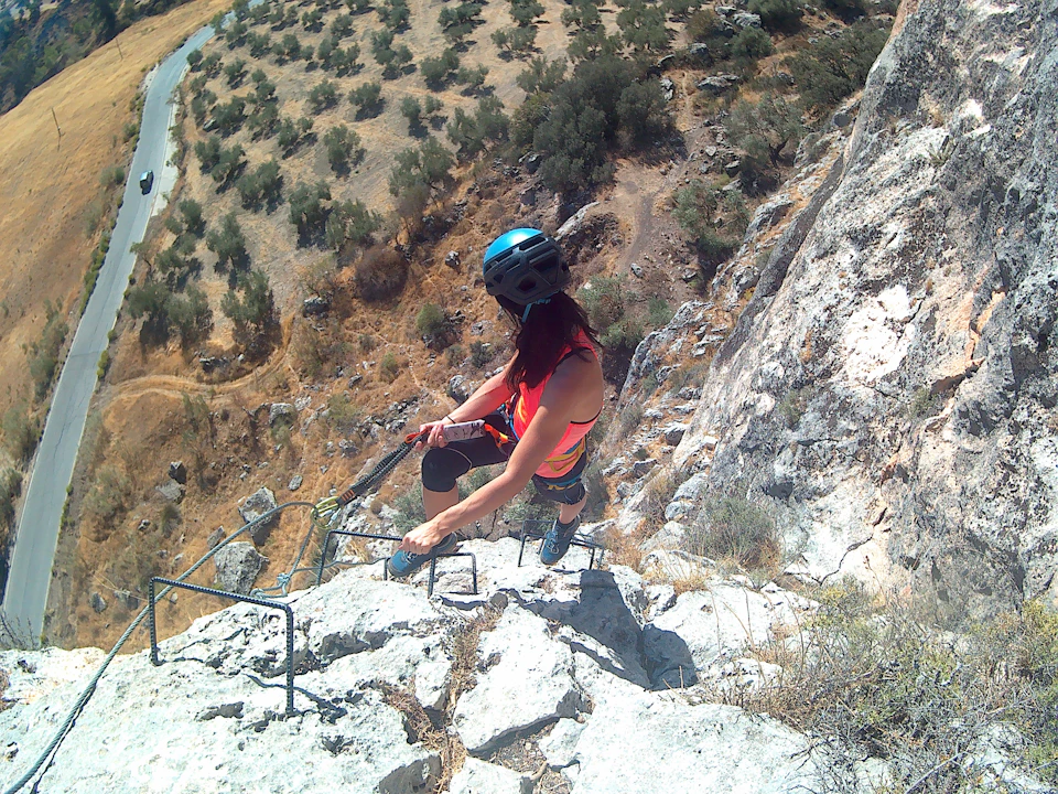A person wearing a helmet climbs a steep rock face using metal rungs, with a road and trees visible below.