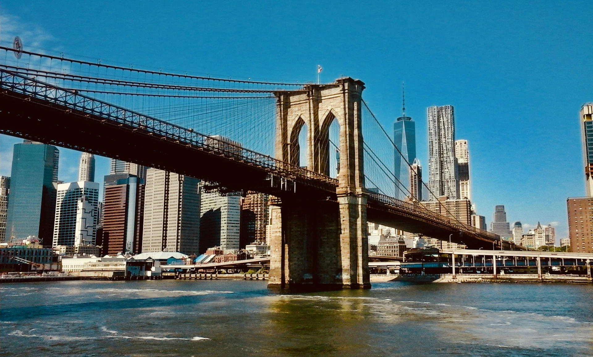 Brooklyn Bridge spans over water with New York City skyline, including One World Trade Center, in the background under a clear blue sky.