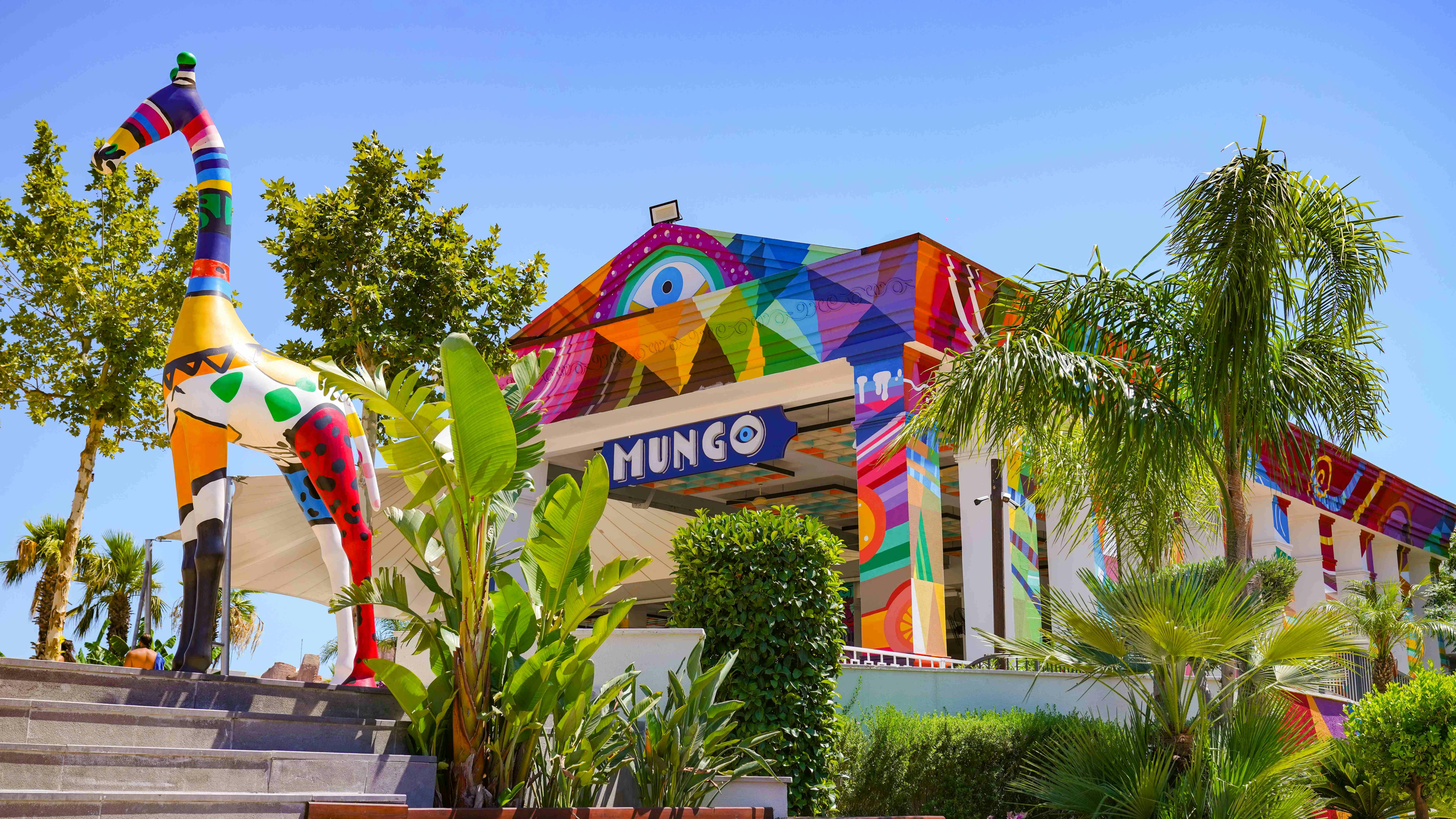 A colorful building named "Mungo" with vibrant geometric patterns, surrounded by greenery under a clear blue sky.