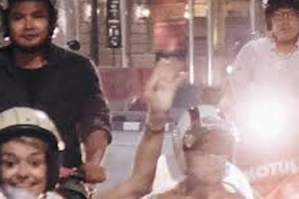 A group of people riding motor scooters with sidecars on a city street at night, buildings and trees in the background.