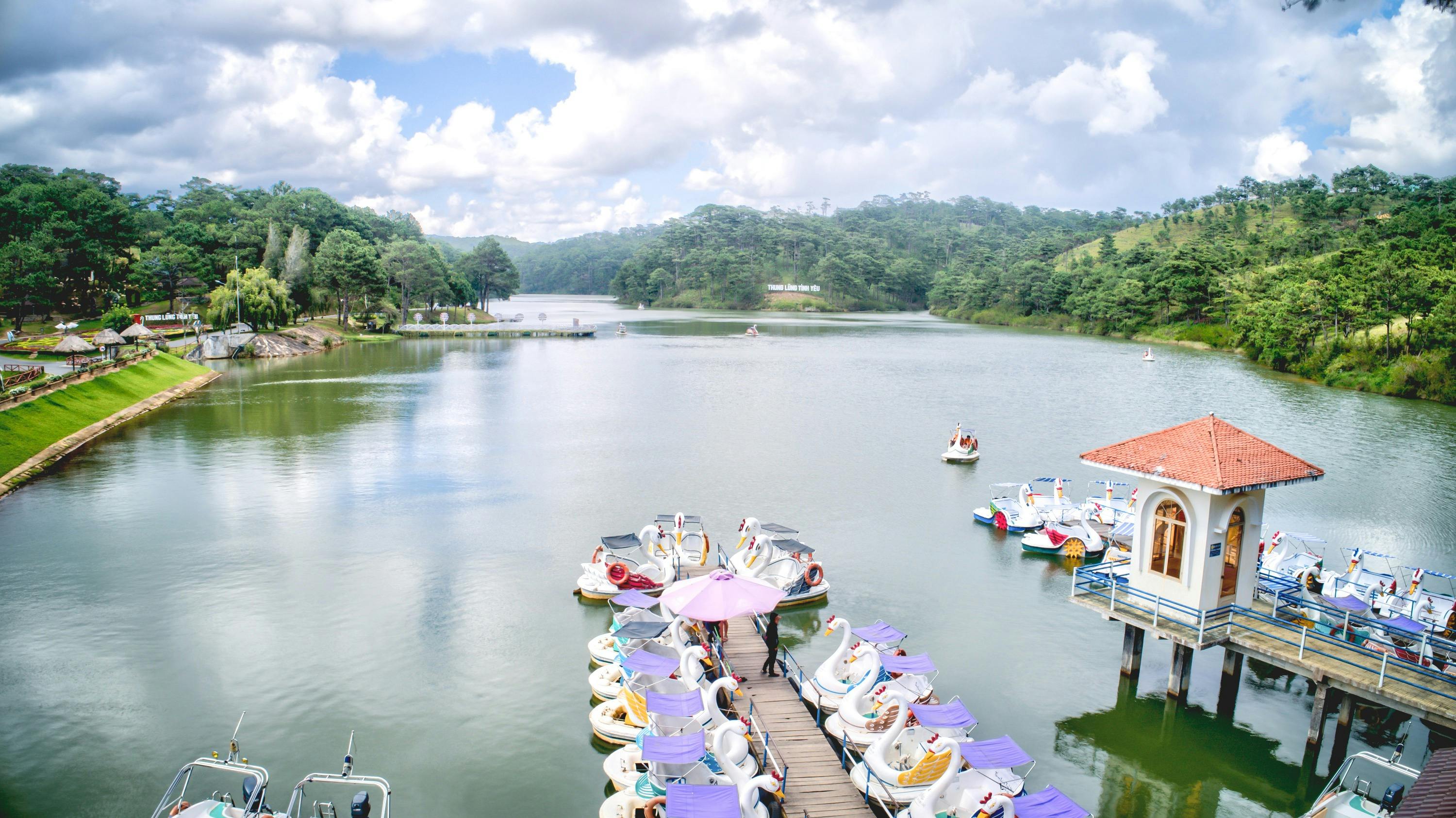 A scenic lake with swan-shaped pedal boats docked by a pier under a pink umbrella, surrounded by trees and hills.