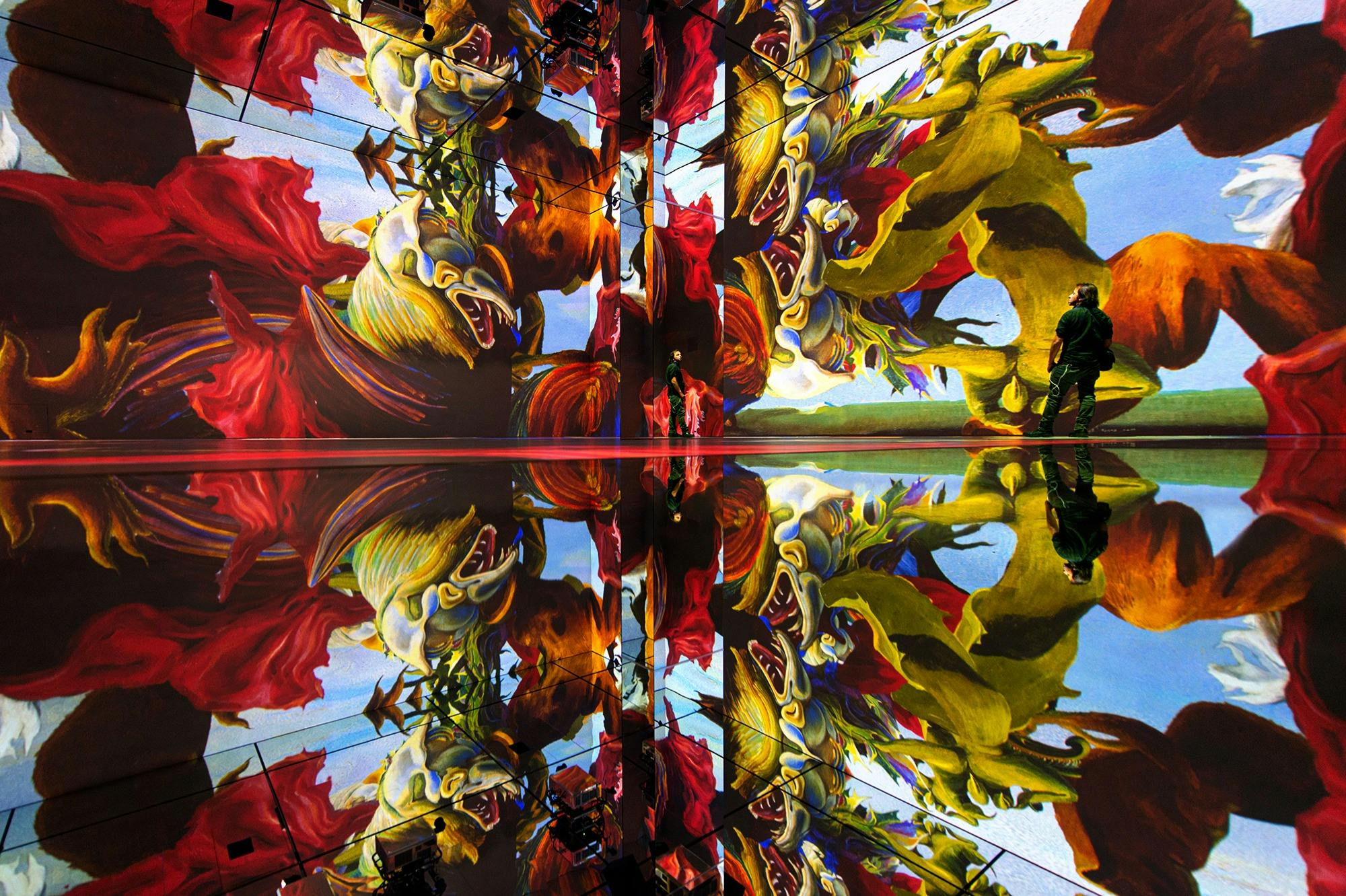 W pomieszczeniu o żywych, lustrzanych projekcjach mitycznych stworzeń i czerwonej draperii stoją dwie osoby.