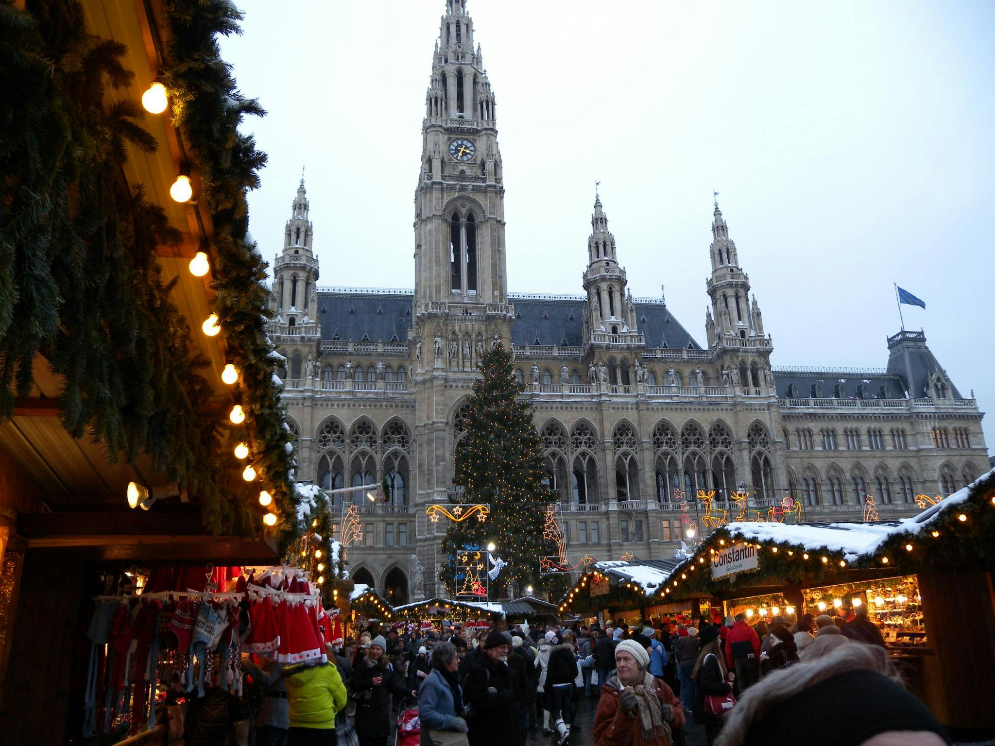 Trångt julmarknad med dekorerade bås, julbelysning och en stor gotisk byggnad i bakgrunden.