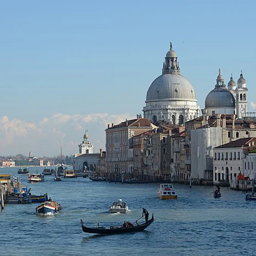 Gondoler och båtar på en kanal i Venedig med historiska byggnader, inklusive välvda strukturer, under en klarblå himmel.
