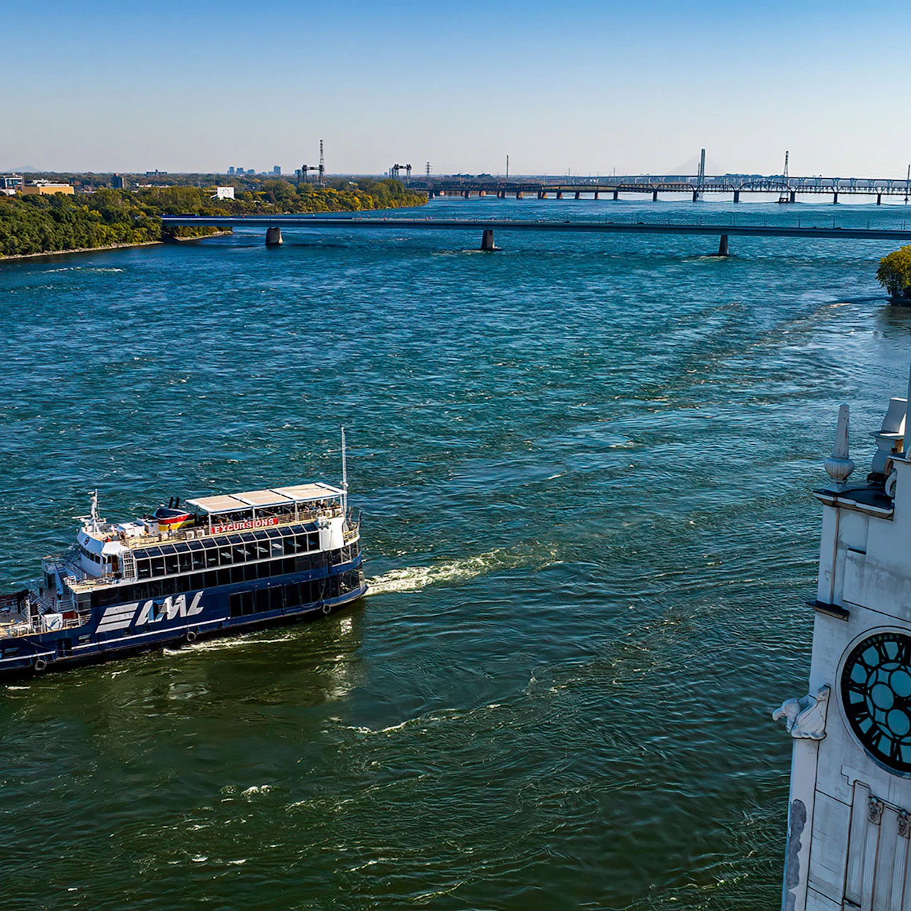 A river with a passenger ferry labeled 'AML' cruising, a bridge in the background, and part of a building with a clock in the foreground.