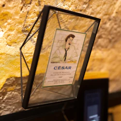 A framed illustration of a man with a bottle, labeled "César," displayed on a stone wall.