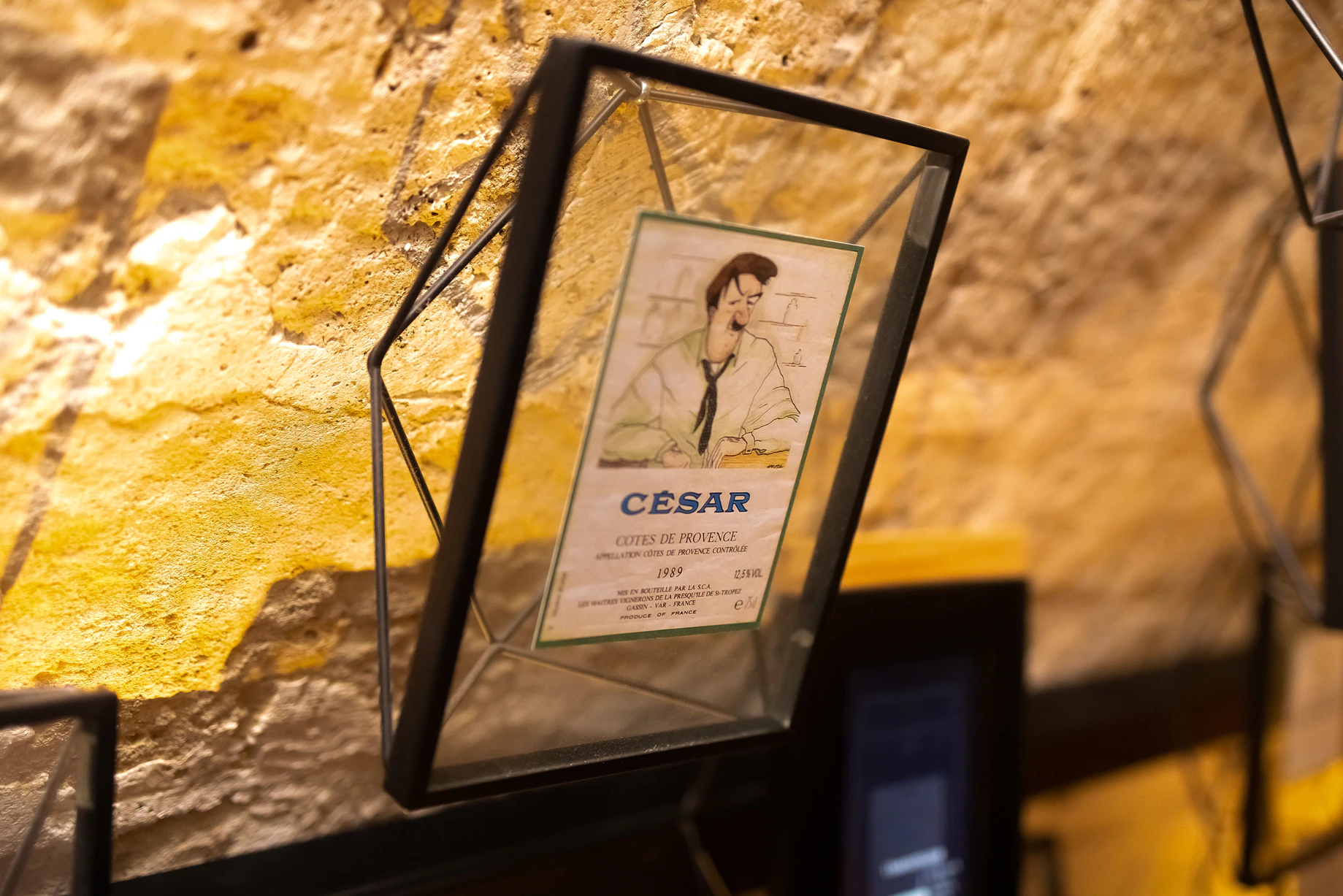 A framed illustration of a man with a bottle, labeled "César," displayed on a stone wall.