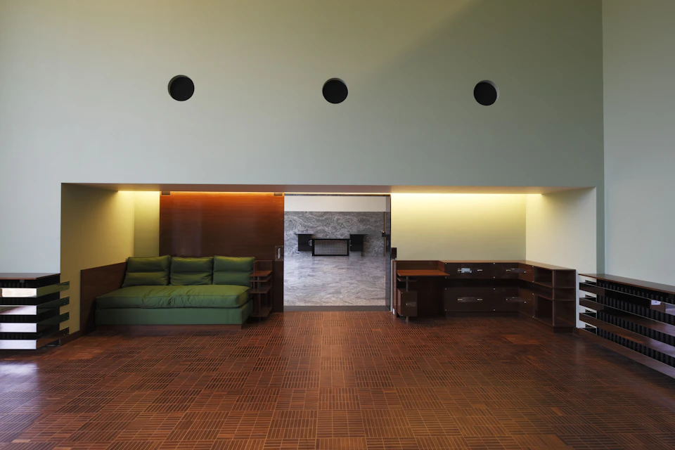 A modern living room with a green sofa, wooden flooring, wall-mounted shelves, and a built-in desk against a marble accent wall.