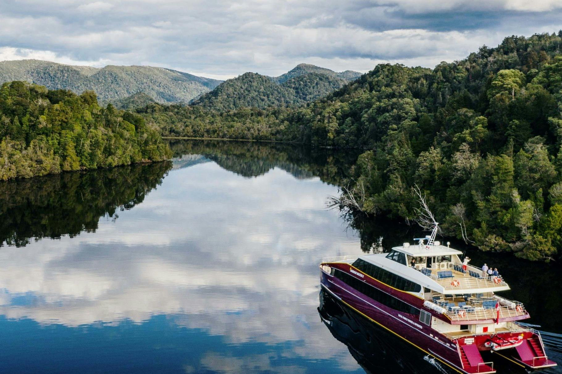 A scenic river surrounded by forested hills, with a maroon and white boat in the foreground on the water.