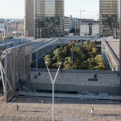 Modern urban plaza med trädäck, träd i en central trädgård och höga glasbyggnader i bakgrunden.