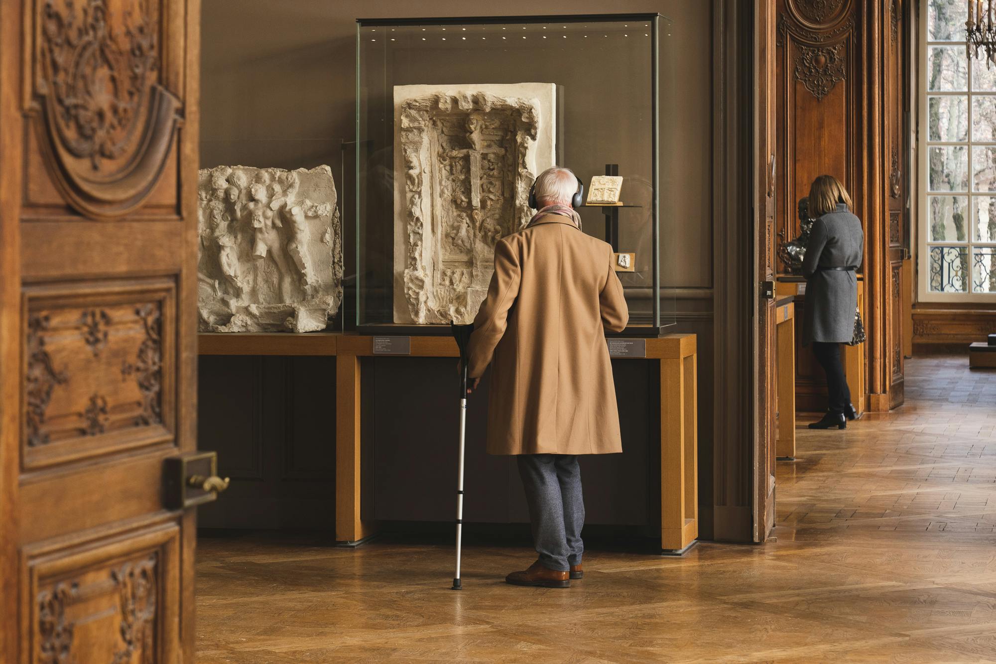 지팡이를 든 노인이 박물관 갤러리의 유리 케이스에 전시된 석고 예술 작품을 감상하고 있습니다.