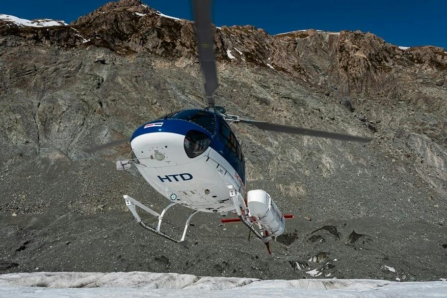 A helicopter flying low over a rocky, mountainous terrain under a clear blue sky.