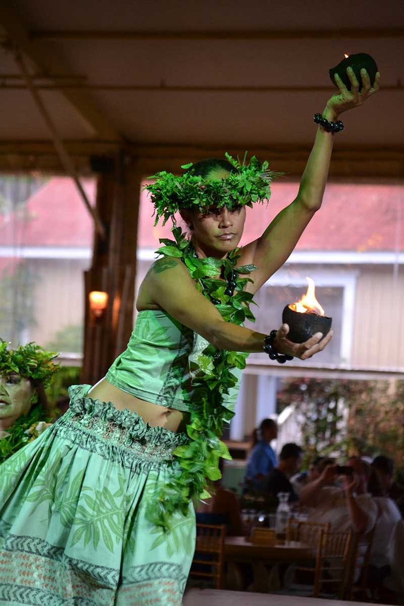 A dancer adorned with leaves and bracelets performs a traditional dance indoors, holding bowls with flames in both hands.