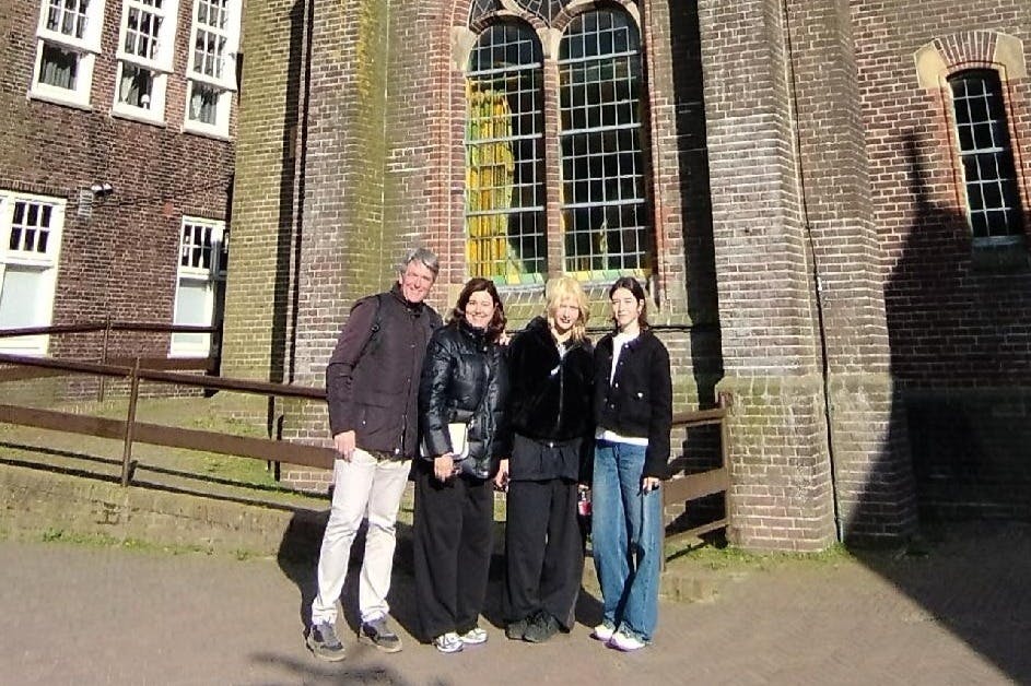 Τέσσερις άνθρωποι στέκονται μπροστά από ένα κτίριο από τούβλα με τοξωτά παράθυρα μια ηλιόλουστη μέρα. Χαμογελούν και ποζάρουν μαζί.