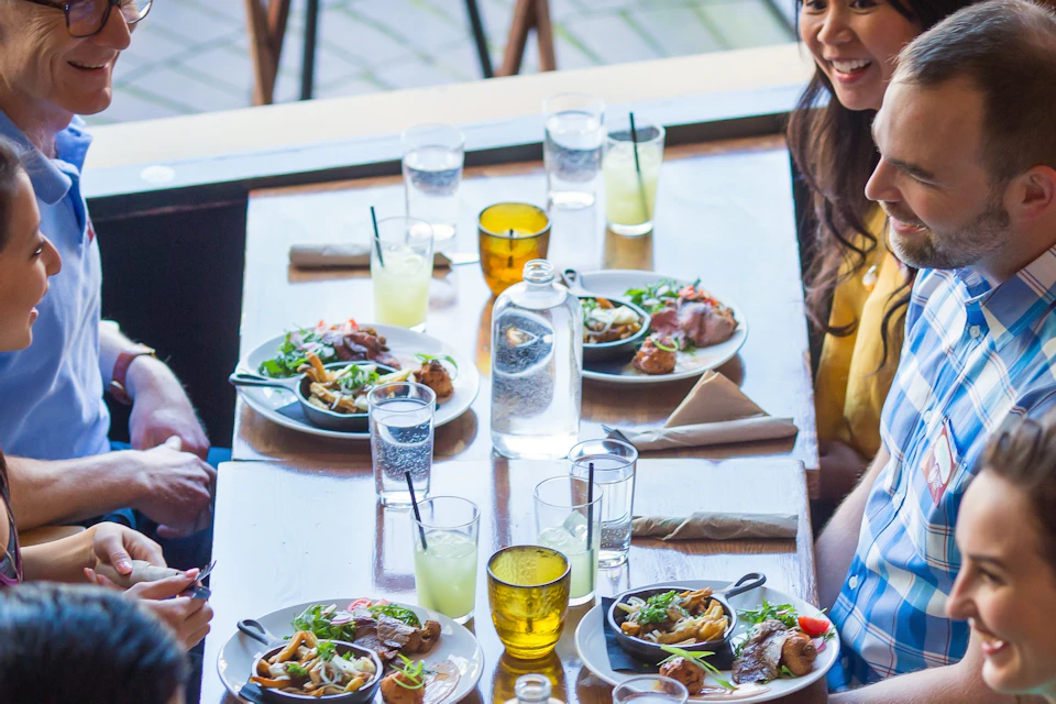 A group of six people sitting around a table sharing a meal with drinks, salads, and plates of food in an outdoor setting.
