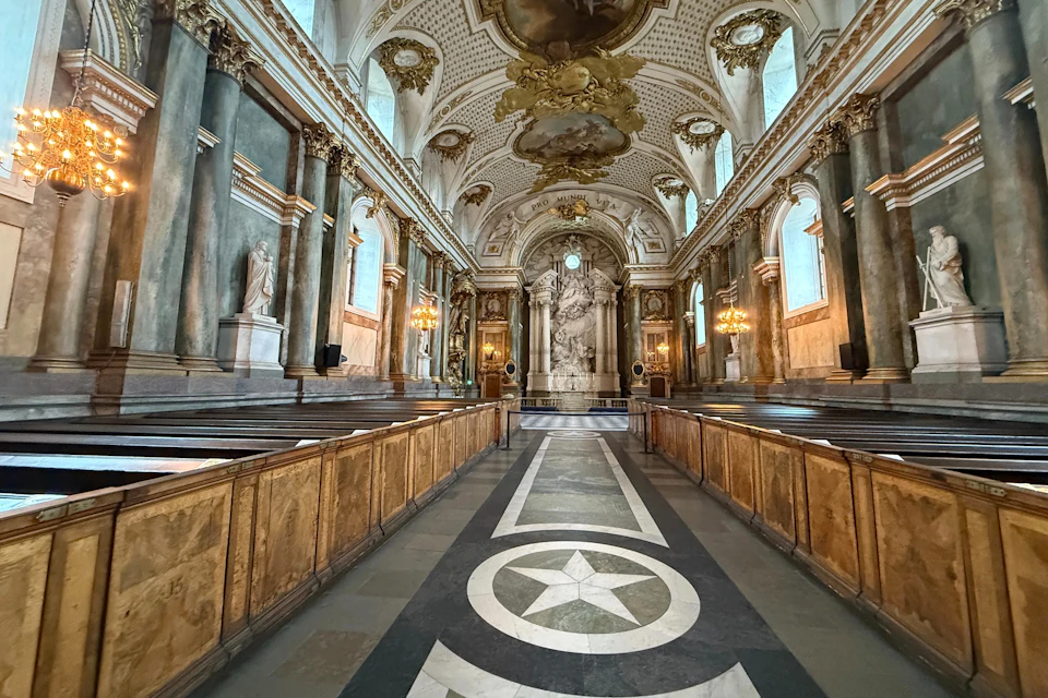 An ornate chapel interior with intricate ceiling designs, grand columns, wooden pews, and a star-patterned floor leading to an altar.