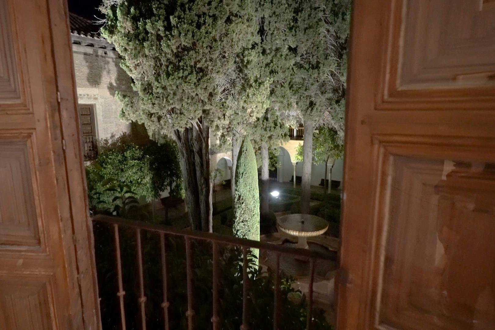 나무 창문 너머로 보이는 높은 나무와 원형 돌 테이블, 아치형 구조물이 있는 안뜰 정원의 야경.
