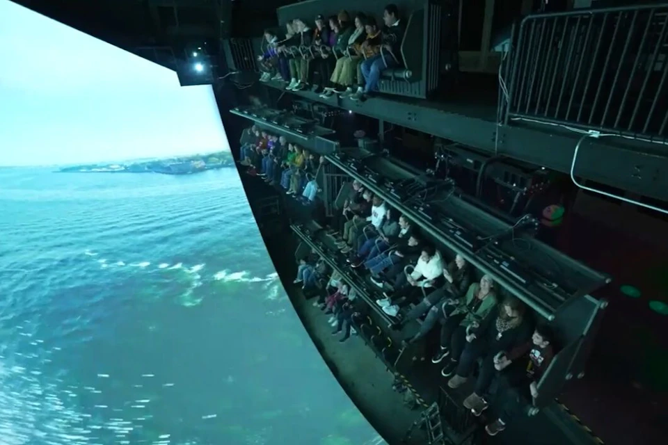 People seated on tiered platforms experiencing a simulator ride with a large curved screen showing an ocean view.