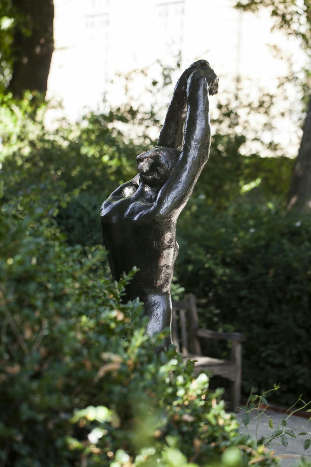 A bronze statue of a nude figure with arms raised overhead, surrounded by lush greenery and a wooden bench in the background.
