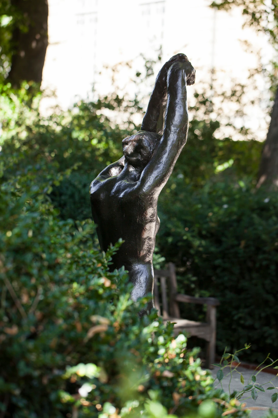 A bronze statue of a nude figure with arms raised overhead, surrounded by lush greenery and a wooden bench in the background.