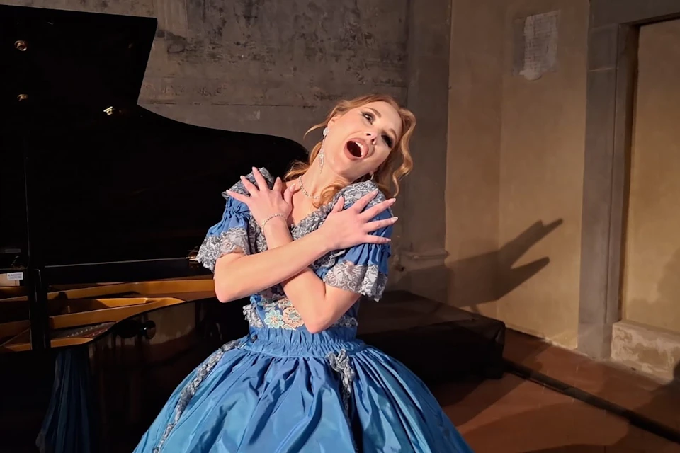 A woman in a blue gown performs dramatically, crossing her arms, in front of a piano in a dimly lit room.