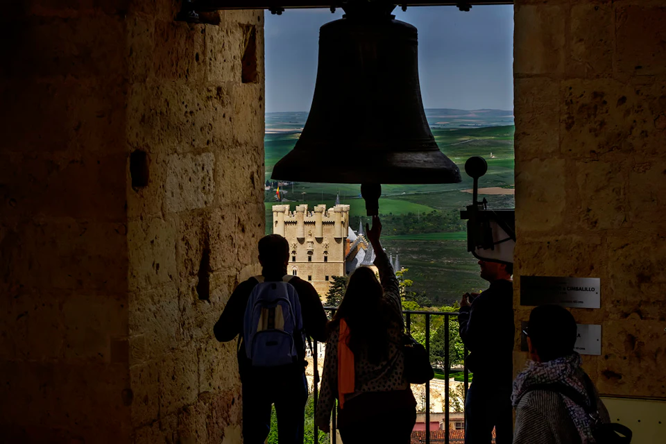 Three people stand under a large bell in a stone tower, looking out at a castle and green landscape in the distance.