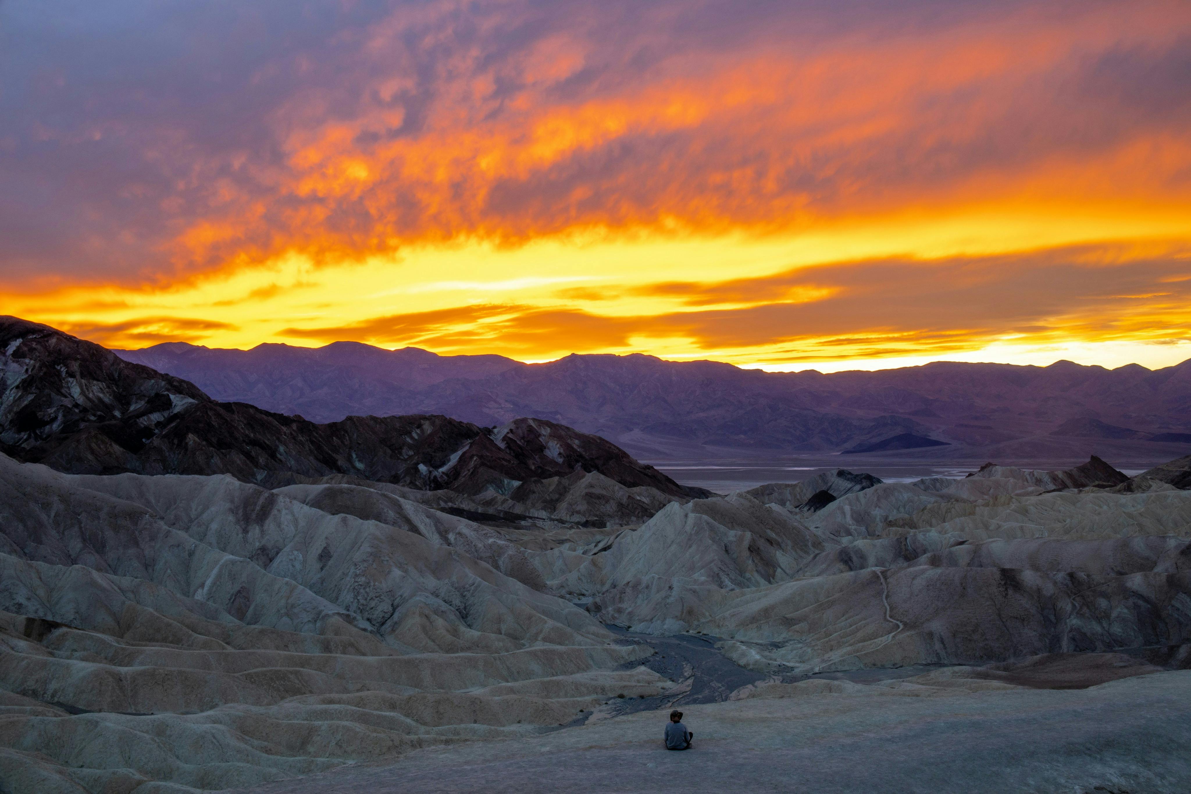 A person sits on a rocky hill at sunrise or sunset, overlooking a desert landscape with colorful clouds in the sky.
