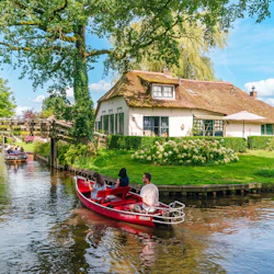 Three people in a red boat on a canal in front of a cottage with a thatched roof, surrounded by greenery and other boats.