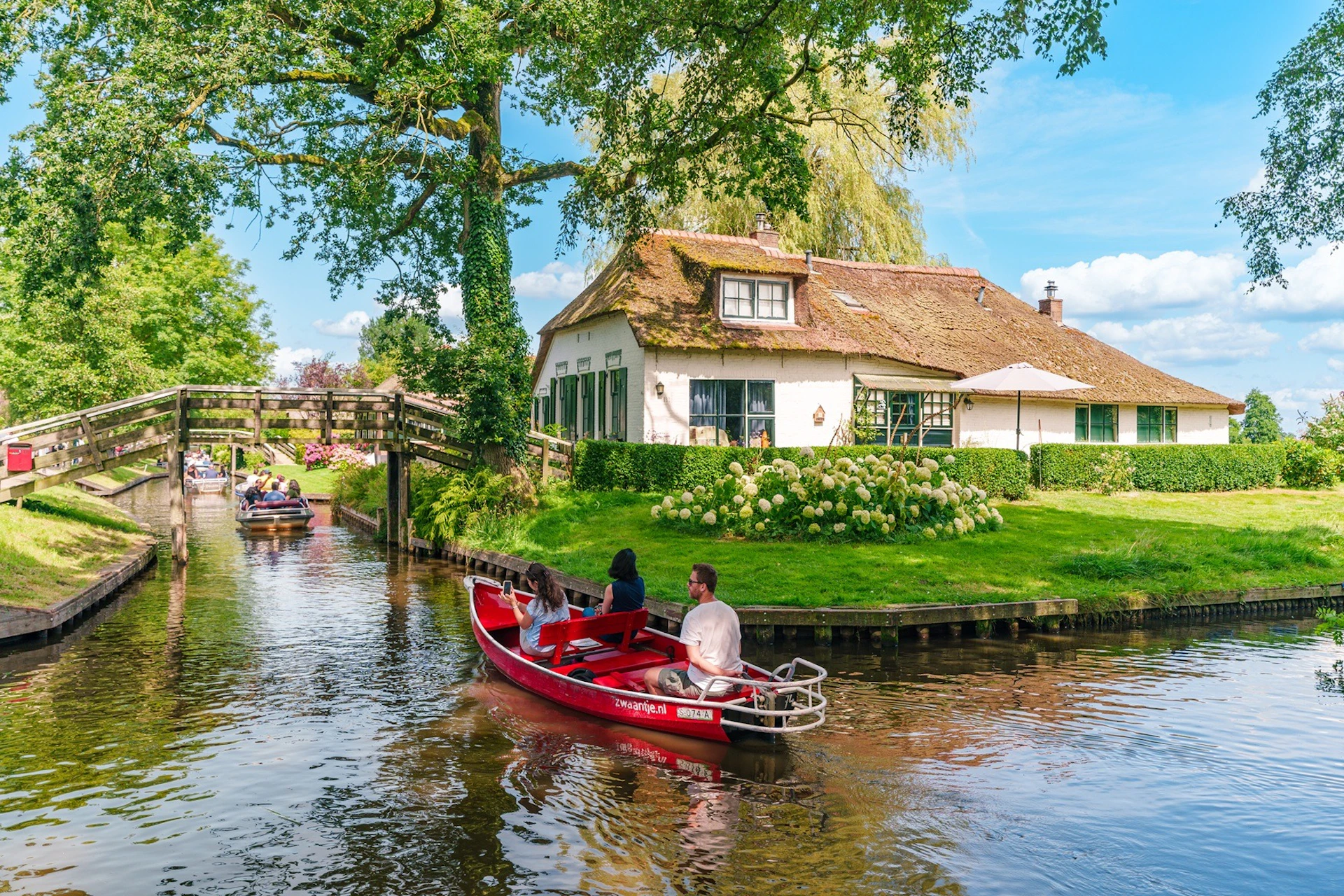 Three people in a red boat on a canal in front of a cottage with a thatched roof, surrounded by greenery and other boats.