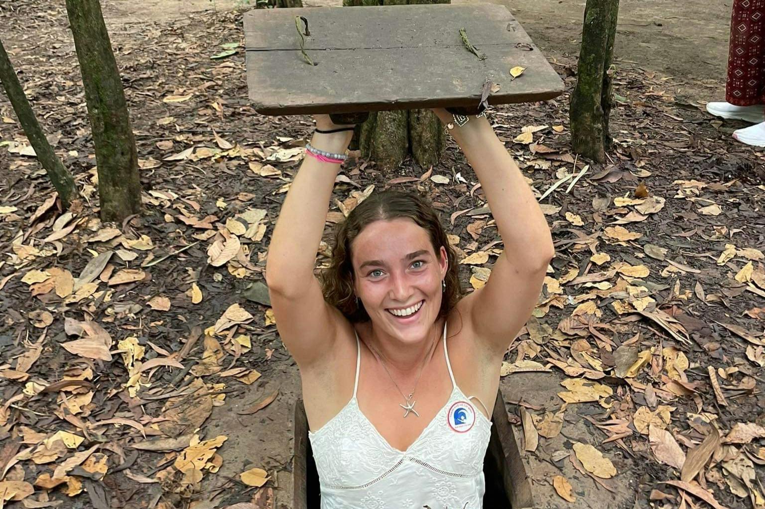 A smiling woman in a white top emerges from a small forest hole, lifting a wooden cover. Fallen leaves surround the area.