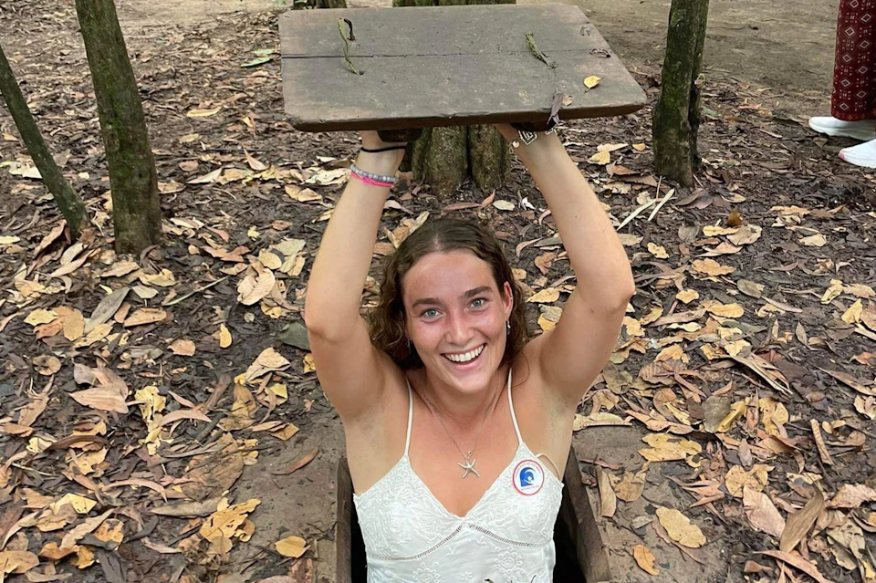 A smiling woman in a white top emerges from a small forest hole, lifting a wooden cover. Fallen leaves surround the area.