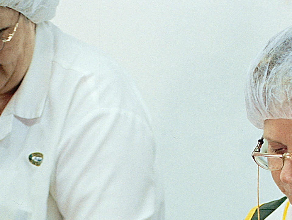 Two workers wearing hairnets, glasses, gloves, and uniforms sorting food items on a conveyor belt in a processing facility.