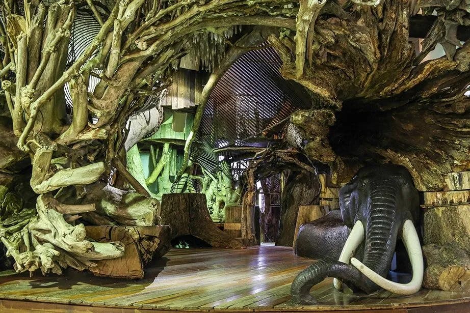 Ένας εσωτερικός χώρος με περίπλοκες ξύλινες και ριζικές δομές, που διαθέτει ένα μεγάλο γλυπτό ελέφαντα και διάφορα καλλιτεχνικά στοιχεία.