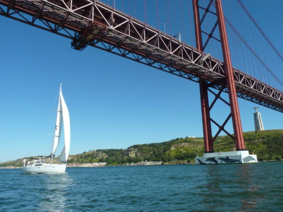 A sailboat on the water passes under a large red suspension bridge with hills and greenery in the background.